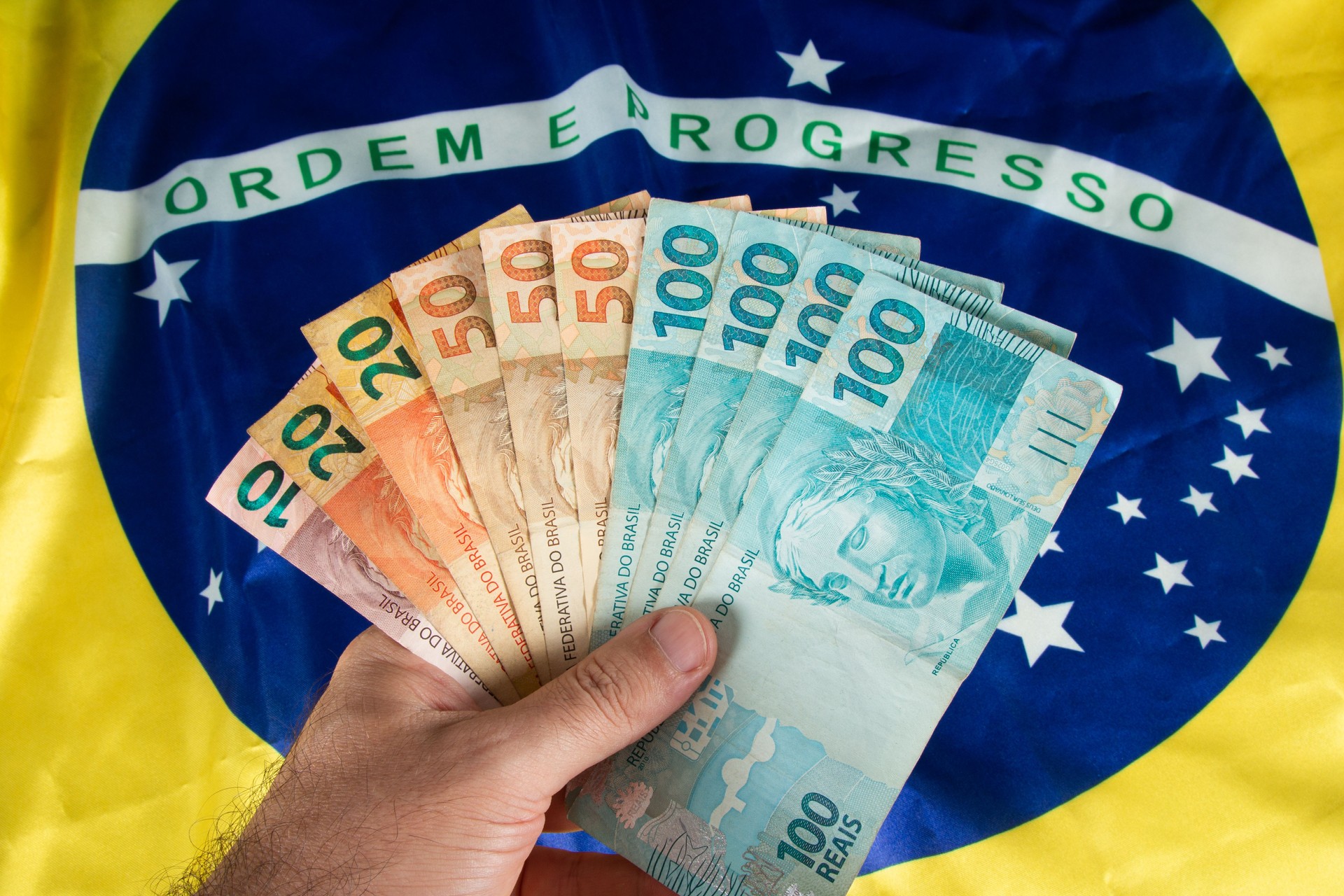 Hand holding Brazilian money banknotes with Brazilian flag in the background
